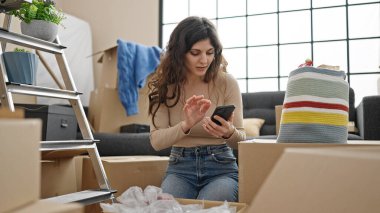 Young beautiful hispanic woman using smartphone unpacking cardboard box at new home