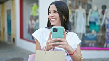 Genç ve güzel İspanyol kadın elbise mağazasında akıllı telefon kullanıyor.