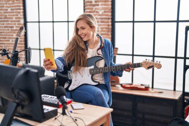 Elektronik gitar çalan genç sarışın müzisyen müzik stüdyosunda akıllı telefondan selfie çekiyor.