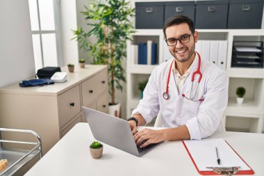 Klinikte çalışan dizüstü bilgisayar kullanan doktor üniforması giyen genç bir İspanyol.