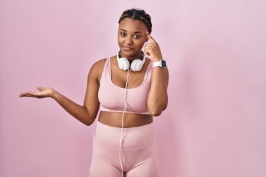 Spor kıyafetleri ve kulaklıkları takan Afro-Amerikan kadın. Kafası karışmış ve avuç içi açık bir şekilde fotokopi alanı göstermekten rahatsız olmuş ve alnını işaret ediyor. Bunu bir düşün.. 
