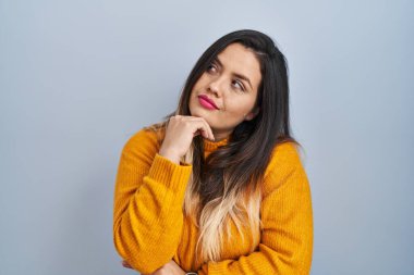 Young hispanic woman standing over isolated background with hand on chin thinking about question, pensive expression. smiling with thoughtful face. doubt concept. 