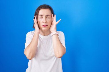 Orta yaşlı Latin bir kadın mavi arka planda, elleri başının üstünde duruyor. Stres yüzünden baş ağrısı çekiyor. migren ağrısı. 