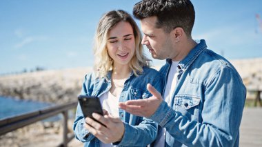 Kadın ve erkek deniz kenarında akıllı telefon kullanarak yan yana duruyorlar.