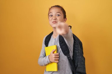 Küçük beyaz çocuk, öğrenci çantası takıyor ve elinde kamerayı işaret eden bir kitap tutuyor. Kendine güvenen bir jest, ciddi görünüyor. 