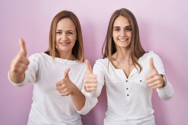 Orta yaşlı anne ve küçük kızı pembe arka planda durup el hareketleriyle olumlu jest yapmayı onaylıyorlar. Gülümsüyorlar ve başarı için mutlular. kazanan jest. 