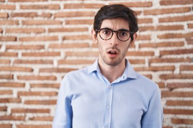 Tuğla duvarın arkasında duran genç İspanyol adam şok olmuş, şüpheci ve alaycı görünüyor, ağzı açıkken şaşırmış. 
