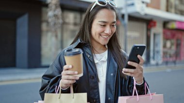 Genç ve güzel İspanyol kadın akıllı telefon kullanıyor. Elinde alışveriş poşetleri ve sokakta kahve tutuyor.