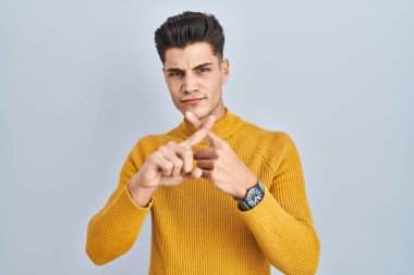 Young hispanic man standing over blue background rejection expression crossing fingers doing negative sign 