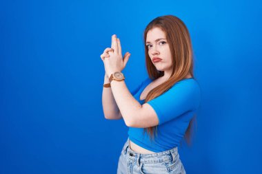 Mavi arka planda duran kızıl saçlı kadın sembolik bir silahla el hareketleriyle cinayet silahlarıyla oynuyor, kızgın surat. 