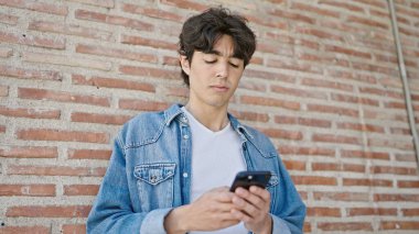 Genç İspanyol adam, izole tuğla arka planda ciddi bir ifadeyle akıllı telefon kullanıyor.