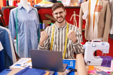 Terzi odasında dizüstü bilgisayar kullanan sakallı İspanyol bir adam çok mutlu ve heyecanlı bir şekilde kolları havada, gülümseyerek ve başarı için bağırarak kazanan bir jest yapıyor. kutlama konsepti. 