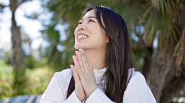 Genç Çinli kadın parkta gökyüzüne bakarak kendinden emin bir şekilde gülümsüyor.