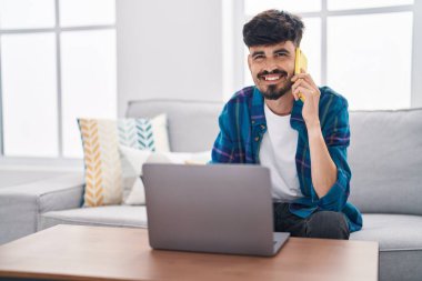 Young hispanic man using laptop and talking on smartphone sitting on sofa at home