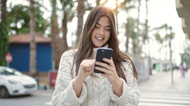 Genç, güzel, İspanyol bir kadın sokakta akıllı telefon kullanarak gülümsüyor.