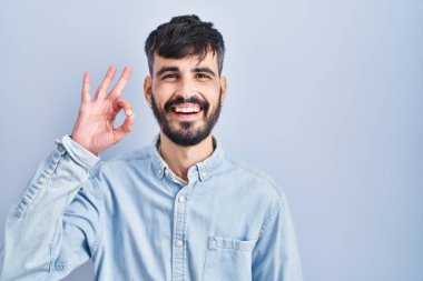 Mavi arka planda sakallı genç İspanyol adam gülümsüyor ve el ve parmaklarıyla 