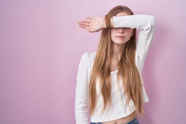 Pembe arka planda duran, kollarıyla gözlerini kapayan, ciddi ve üzgün görünen beyaz bir kadın. Görme, saklama ve reddetme konsepti 