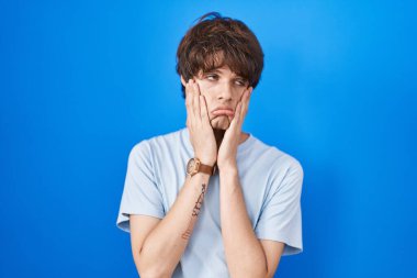 Hispanic young man standing over blue background tired hands covering face, depression and sadness, upset and irritated for problem 