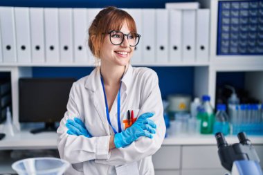 Genç bilim kadını gülümsüyor. Kollarını kavuşturup laboratuvarda duruyor.