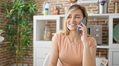 Genç sarışın kadın yemek odasında masada oturmuş akıllı telefondan konuşuyor.