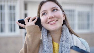 Akıllı telefonun yanındaki sesli mesajı dinleyen genç sarışın kadın sokakta kahve içiyor.