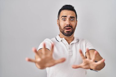 Beyaz arka plan üzerine gündelik giysiler giymiş sakallı genç bir İspanyol. Korkudan dehşete düşmüş ve korkmuş. El hareketlerini durdur, şok içinde bağır. panik konsepti. 