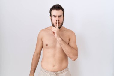 Beyaz arka planda üstsüz duran İspanyol genç adam sessiz olmak istiyor. sessizlik ve gizli konsept. 