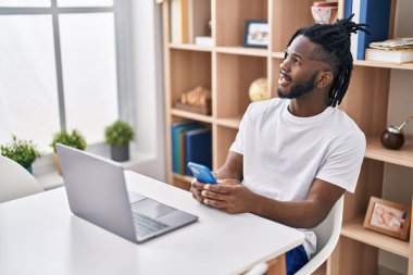 Evde masada oturan, dizüstü bilgisayar ve akıllı telefon kullanan Afrikalı Amerikalı bir kadın.