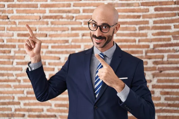 秃头的男人 留着胡子 穿着商务服装 戴着眼镜 微笑着看着摄像机 用双手和手指指向旁边 — 图库照片