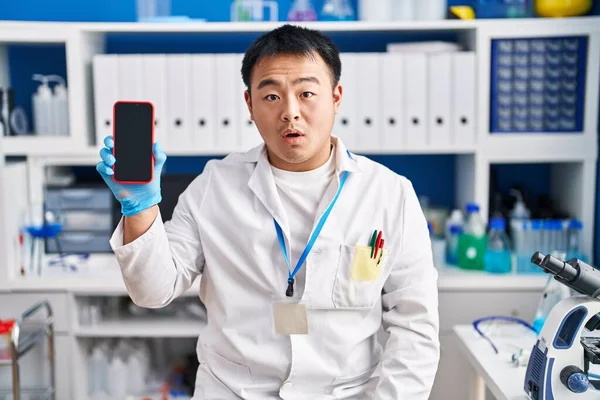 Young chinese man working at scientist laboratory holding smartphone scared and amazed with open mouth for surprise, disbelief face