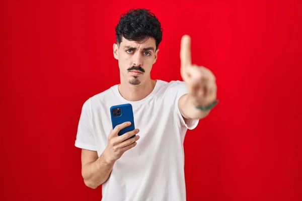 Латиноамериканец Использует Смартфон Красном Фоне Показывая Пальцем Вверх Злое Выражение — стоковое фото