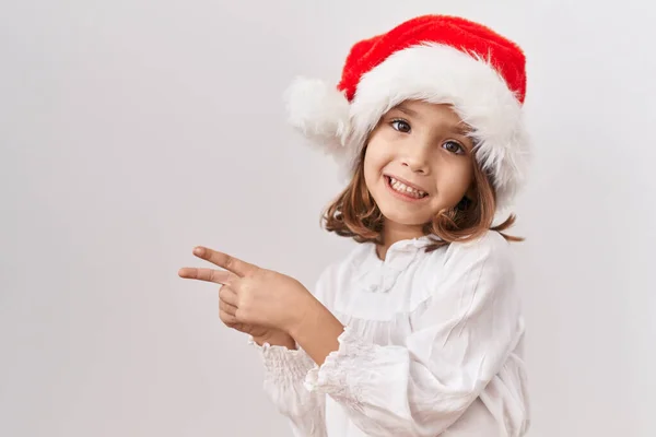 他那头戴圣诞礼帽的惊慌失措的小女孩笑着看着摄像机 用双手和手指指向旁边 — 图库照片