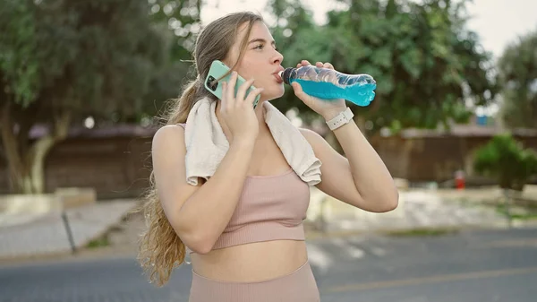 年轻的金发女子穿着运动服 在公园里喝着精力充沛的饮料 用智能手机交谈 — 图库照片