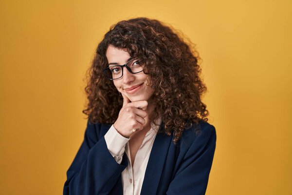 Hispanic woman with curly hair standing over yellow background looking confident at the camera smiling with crossed arms and hand raised on chin. thinking positive. 