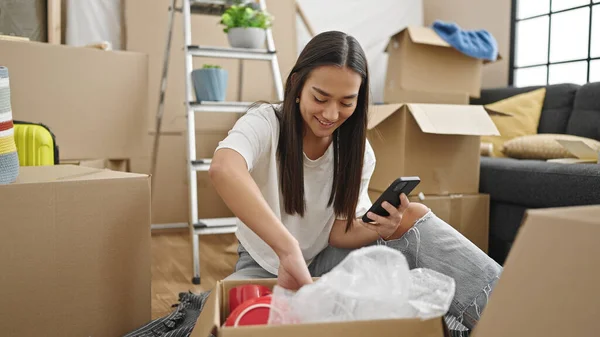 Young beautiful hispanic woman unpacking cardboard box using smartphone at new home