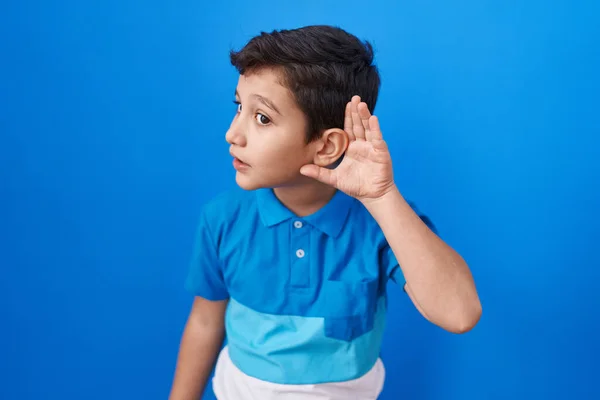 小さなヒスパニック系の少年の青い背景に手で耳を傾け 噂やゴシップに耳を傾け笑みを浮かべて立っている 聴覚障害の概念 — ストック写真