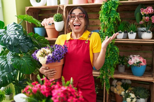 Young arab woman working at florist shop holding plant celebrating victory with happy smile and winner expression with raised hands