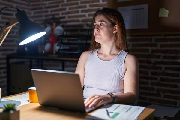 Brunette woman working at the office at night smiling looking to the side and staring away thinking.