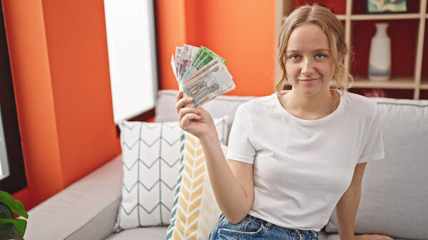Молодая блондинка улыбается уверенно держа дома банкноты российских рублей