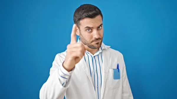 年轻的惊慌失措的医生站在那里 脸上带着不高兴的表情 用手指对着相机在孤立的蓝色背景上指指点点 — 图库照片