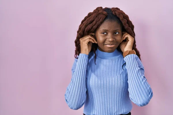 ピンクの背景に立っているアフリカの女性は 大きな音楽のノイズに悩まされた表情で指で耳を覆います 聴覚障害の概念 — ストック写真