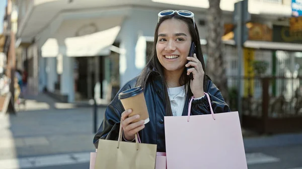 若い美しいヒスパニック系の女性がコーヒーショップテラスでショッピングバッグやコーヒーを保持しているスマートフォンで話しています — ストック写真