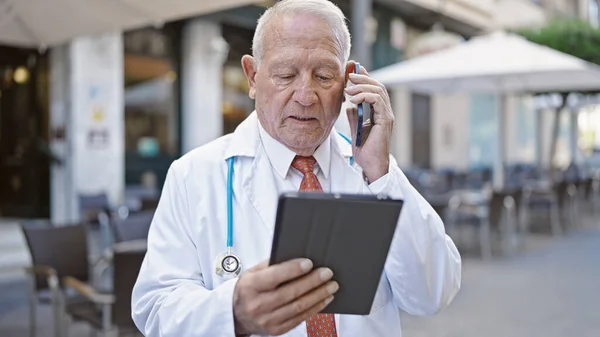 コーヒーショップのテラスでスマートフォンで話すタッチパッドを使用してシニア白髪の男性医師 — ストック写真