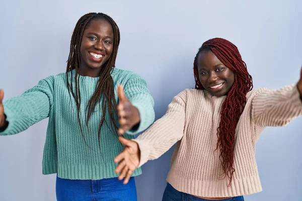 两个非洲女人站在蓝色的背景上 看着摄像机 张开双臂拥抱 欢快的表达拥抱幸福 — 图库照片