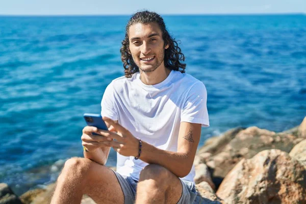 Young Hispanic Man Using Smartphone Sitting Rock Seaside — Stockfoto