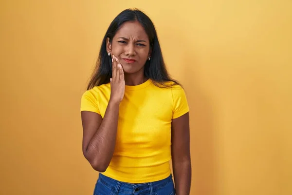 Young indian woman standing over yellow background touching mouth with hand with painful expression because of toothache or dental illness on teeth. dentist