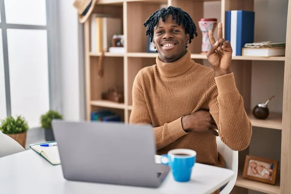 コンピュータのラップトップを使用して働いている恐ろしいロックを持つ若いアフリカの男は 勝利の兆候を行うカメラで幸せな顔をウィンクで笑っています — ストック写真