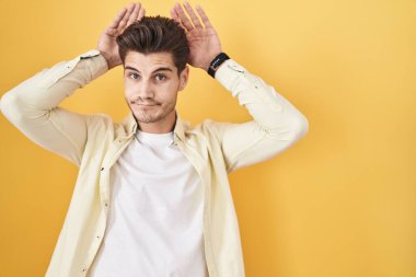 Young hispanic man standing over yellow background doing bunny ears gesture with hands palms looking cynical and skeptical. easter rabbit concept. 