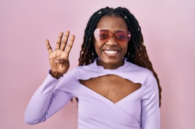 Pembe arka planda örülü saçları olan Afrikalı bir kadın dört numarayı gösterip işaret ederken kendinden emin ve mutlu gülümsüyor.. 