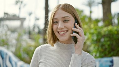 Genç sarışın kadın gülümsüyor. Parkta akıllı telefondan konuşuyor.